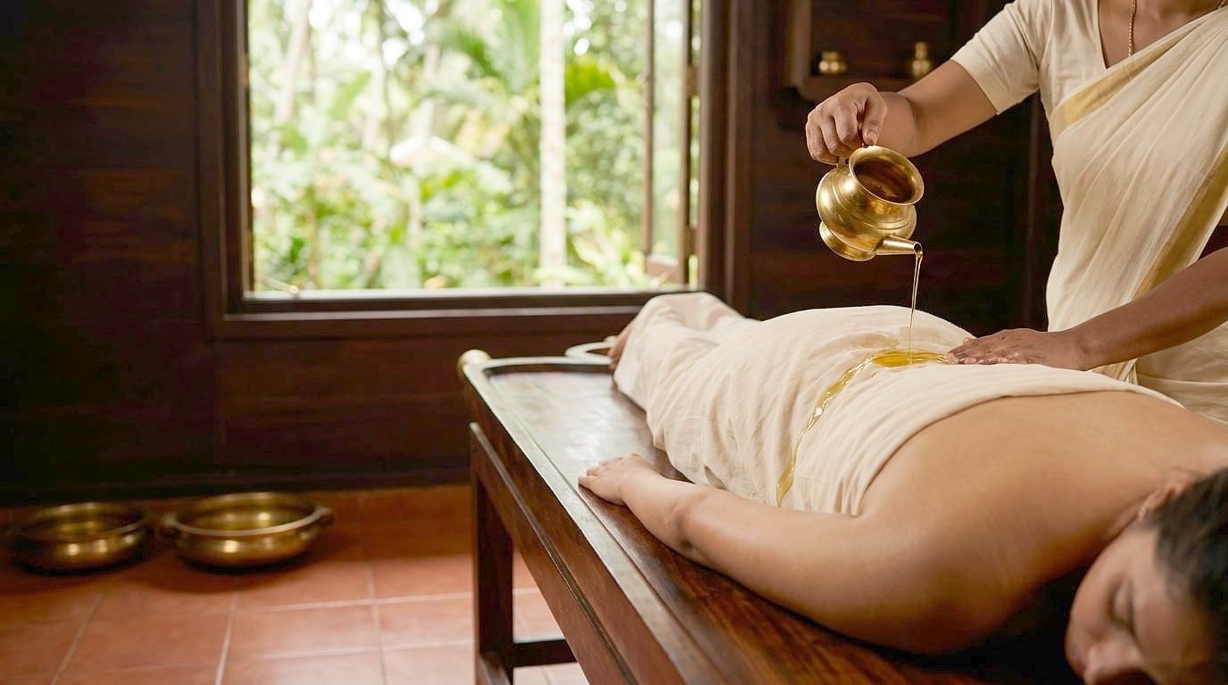 A therapist pours warm oil from a brass vessel onto the back of a person lying on a wooden table