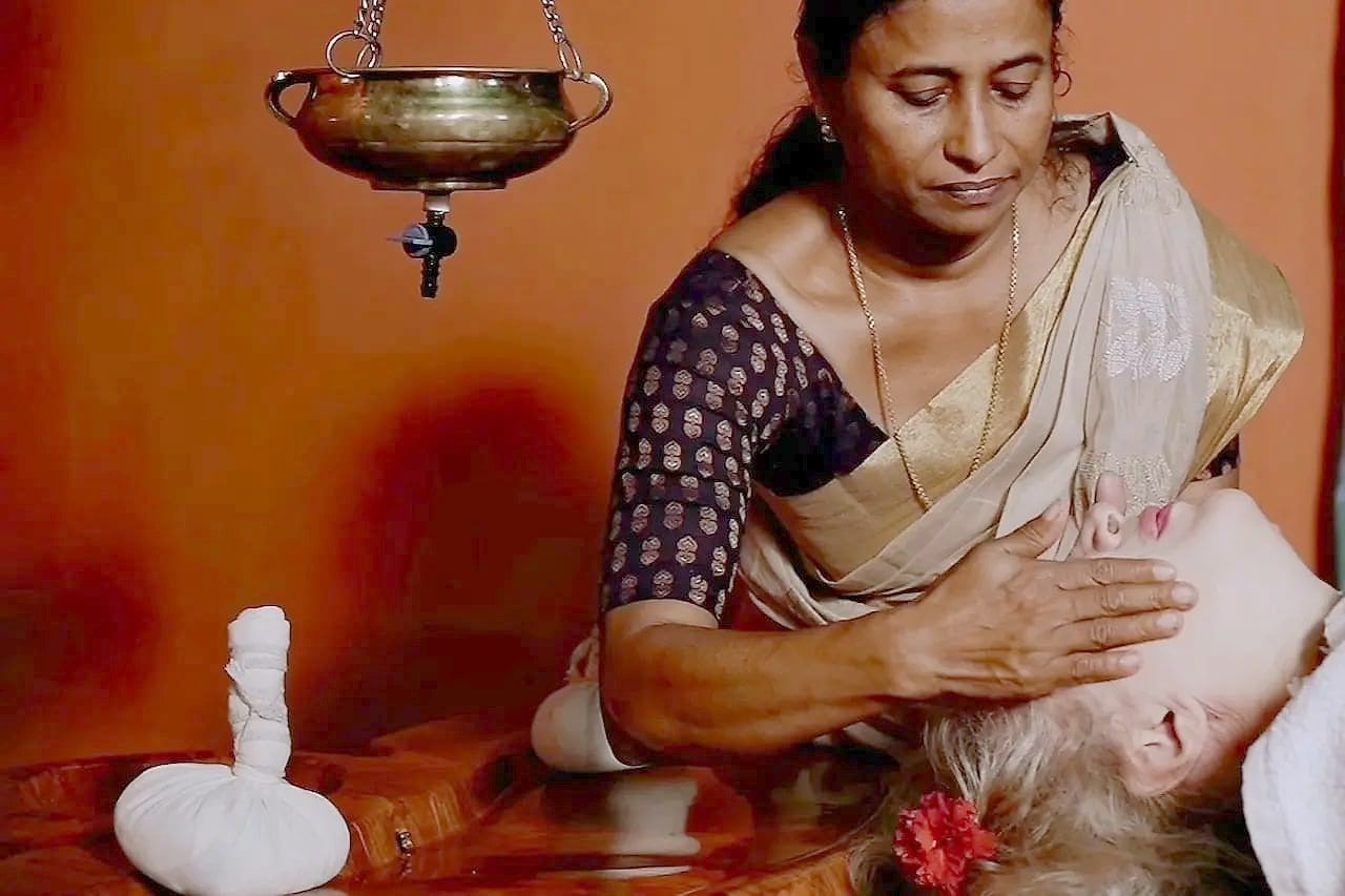 A therapist giving a head and face massage in a treatment room, a brass shirodhara vessel above the table and herbal boluses nearby